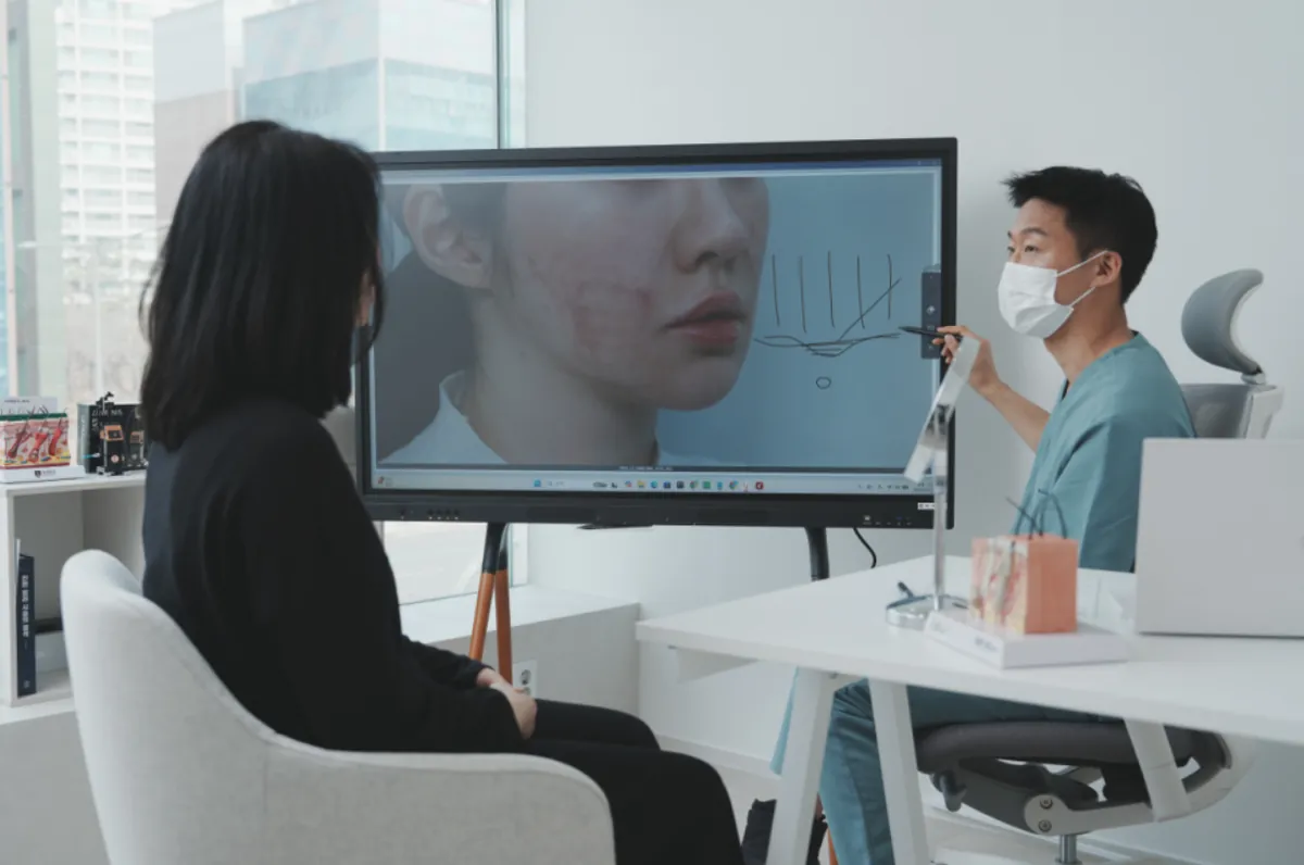 A doctor at a Jamsil dermatology clinic pointing to a skin photo on a digital whiteboard while explaining to a patient